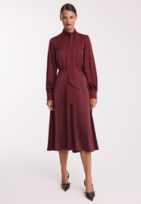 Burgundowa sukienka z wysokim kołnierzem, długimi rękawami i wiązaniem przy szyi. Gładka tekstura, lekko opływający fason, długość do kolan z rozkloszowanym dołem.