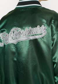 Grüne Satin-Bomberjacke mit einem gerippten Kragen und dem "Huf Worldwide"-Logo in Weiß und Silber auf dem Rücken. Glatte Textur, glänzende Oberfläche.