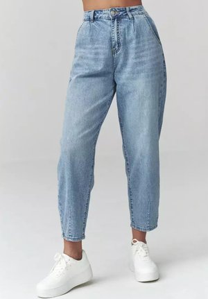 Jasnoniebieskie dżinsy z wysokim stanem, zwężane nogawki, noszone przez stojącą osobę wraz z białymi sneakersami na platformie.