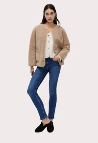 Beige sherpa-jacka med rundad krage, två framfickor, knäppning med knappar, vit ribbad topp, blåa smala jeans och svarta slip-on skor.