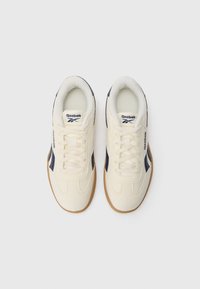 Krēma krāsas kedas ar tumši ziliem akcentiem, ar noapaļotu formu, auduma augšdaļu un gumijas zoli. Šņorēšanas auklas un Reebok logo uz mēles.
