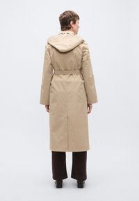 Μπεζ μακρύ παλτό με κουκούλα, ζώνη στη μέση και ευθύ κόψιμο. Το υλικό φαίνεται λείο; διαθέτει κεντρικό πίσω σκίσιμο για ευκολία στην κίνηση.