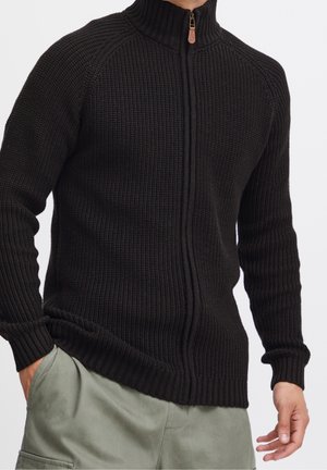 Mężczyzna w czarnym dzianinowym swetrze na zamek z wysokim kołnierzem i jasnoszarych spodniach, dłonie swobodnie opuszczone wzdłuż ciała na białym tle.