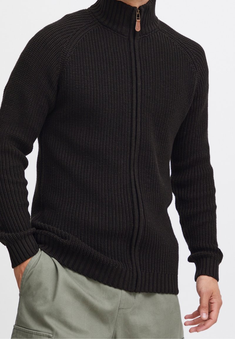 Homme portant un pull en maille noir à fermeture éclair avec col montant et pantalon gris clair, les mains reposant le long du corps sur un fond blanc.