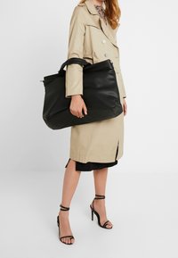 Černá kožená taška přes rameno s texturovaným povrchem, velká velikost, dvojité horní držadla a stříbrné kovové detaily, kterou drží osoba v béžovém kabátu.