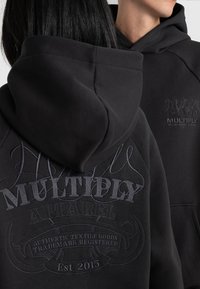 Black hooded sweatshirt featuring textured embroidery with "MULTIPLY APPAREL" and details. Smooth fabric, relaxed fit, and kangaroo pocket.