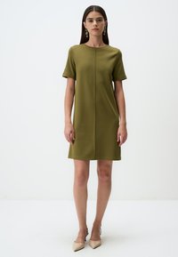 Olivengrünes Kurzarmkleid mit geradem Schnitt, strukturiertem Stoff und Mittelnaht-Detail. Getragen mit hellen, spitzen Absätzen.