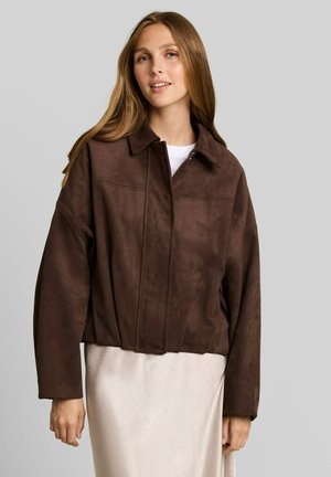 Giovane donna con capelli lunghi indossa una giacca in camoscio marrone con cerniera sopra una maglietta bianca e una gonna beige chiaro, in piedi davanti a uno sfondo grigio uniforme.