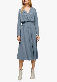 Kobieta w długiej, niebiesko-szarej sukience midi z długim rękawem i zebranym w talii paskiem, w beżowych botkach do kostek, stojąca na białym tle.