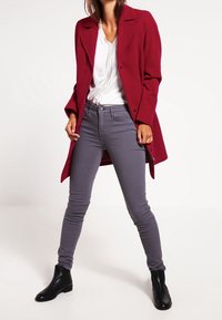 Burgundský kabát přes bílou blůzu, kombinovaný se šedými skinny džíny a černými kotníkovými botami. Oblečení se vyznačuje čistými liniemi a přiléhavým střihem.
