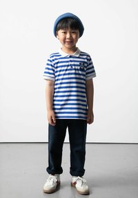 Pasiakowana niebiesko-biała koszulka polo z kołnierzykiem i kieszonką, granatowe spodnie i białe tenisówki z czerwonymi akcentami. Niebieska czapka na głowie.