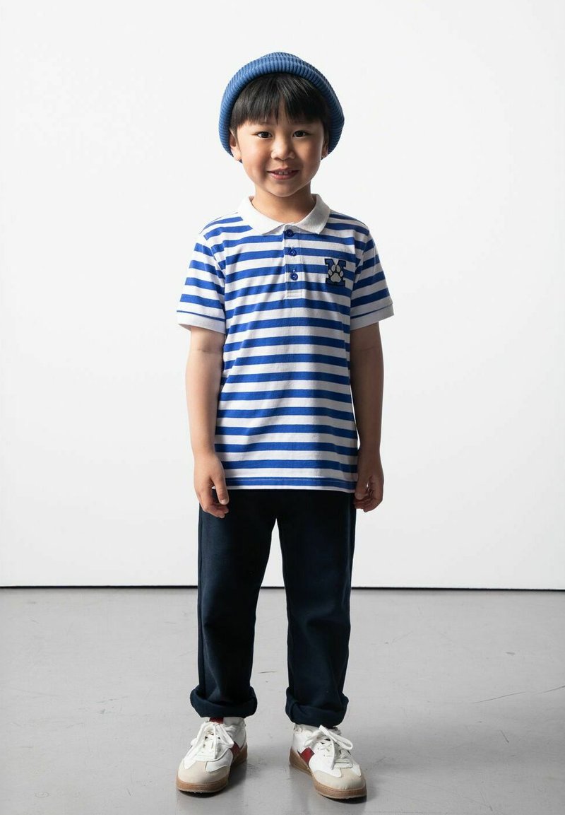 Pasiakowana niebiesko-biała koszulka polo z kołnierzykiem i kieszonką, granatowe spodnie i białe tenisówki z czerwonymi akcentami. Niebieska czapka na głowie.