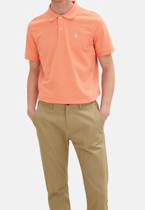 Mężczyzna w łososiowej koszulce polo z krótkim rękawem i beżowych spodniach, z rękami w kieszeniach, na jednolitym tle.