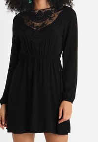 Vestido negro con mangas largas y cintura elástica. Presenta un yugo de encaje transparente con bordados florales y un corte holgado.
