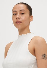 Hvit, ribbestrikket ermeløs topp med høy hals og volangkant. Har en tettsittende design og glatt tekstur, kombinert med hoops-øreringer.