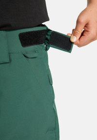 Groene stoffen sneeuwbroek met verstelbare tailleband en een zwarte klittenbandsluiting. Zijzak zichtbaar nabij de tailleband.