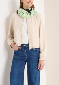 Giacca bomber beige con zip, polsini elasticizzati e girovita regolabile, abbinata a jeans blu a gamba larga e una sciarpa verde a fantasia.