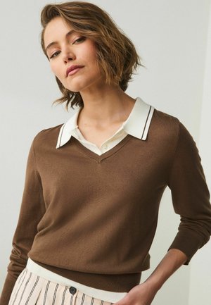 Woman with short brown hair wears a brown V-neck sweater over a white collared shirt and beige striped pants, standing against a plain background.