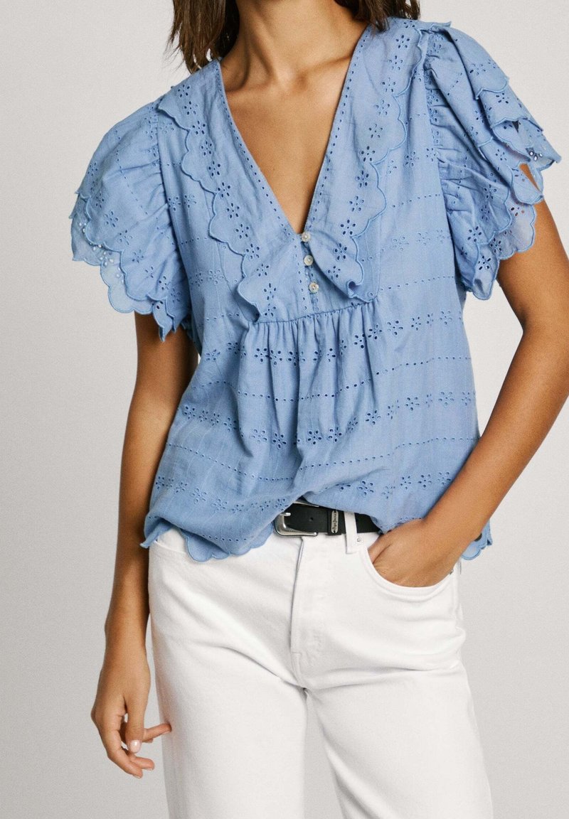Blusa azul claro con mangas onduladas, detalles de ojal y un escote en V profundo. Cuenta con botones en la parte delantera. Combinada con pantalones blancos.