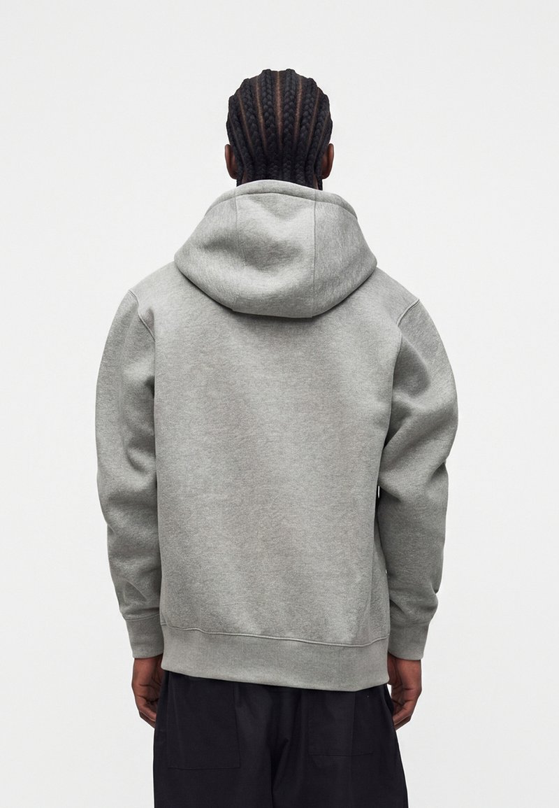 Persona con el pelo trenzado que lleva una sudadera con capucha lisa de color gris claro y pantalones negros, vista desde atrás sobre un fondo blanco.