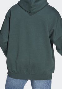 Person klädd i en mörkgrön oversized hoodie med huva och ljusblå jeans, sedd bakifrån mot en neutral bakgrund.