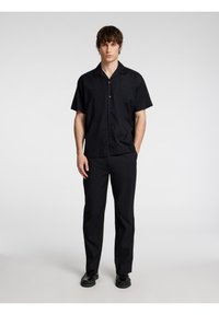 Camicia nera a maniche corte con colletto, chiusura a bottoni e tessuto liscio abbinata a pantaloni neri e scarpe nere. Look semplice e coerente.