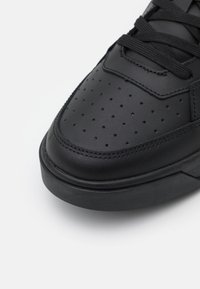 Bout de basket en cuir noir avec perforations, lacets noirs et coutures visibles sur fond blanc.
