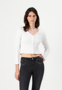 Cardigan corto blanco con relieve, de mangas largas y cuello en V, con seis botones en la parte frontal. Combinado con jeans de mezclilla negros decorados con tachuelas.