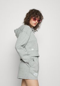 Sweatshirt cinza cortada com cordão e logótipo, combinada com calções iguais que incluem bolsos laterais e logótipo. Usado com óculos de sol cor-de-rosa.