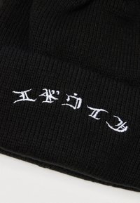 Gorro preto canelado com texto bordado em branco. O material é malha elástica, proporcionando calor e conforto. Design simples sem enfeites.