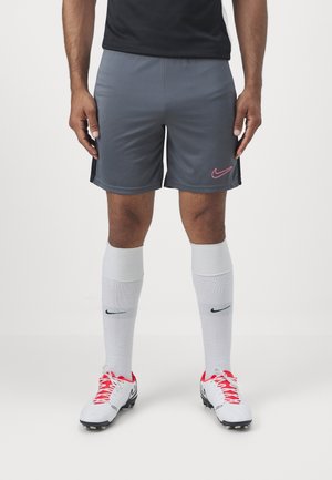 Homme portant un short de sport gris Nike, des chaussettes blanches Nike montantes, des crampons blancs avec des lacets rouges, debout sur une surface blanche.