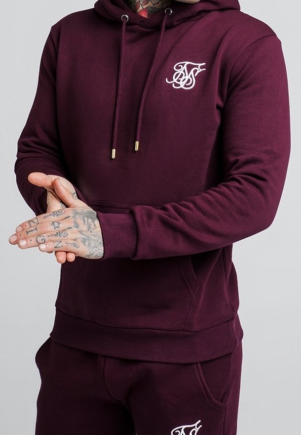 Homme portant un sweat à capuche bordeaux et un pantalon assorti avec un logo blanc brodé, mains jointes devant, tatouages visibles sur les mains et le cou.