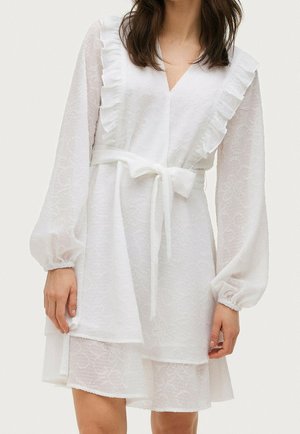 Kobieta w białej, fakturowanej sukience z długimi bufkowatymi rękawami, marszczonymi ramionami, dekoltem w serek i przewiązanym paskiem w talii.