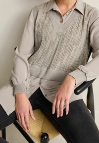 Gilet grigio chiaro in maglia indossato sopra una blusa beige a maniche lunghe, con polsini a coste e scollatura a V, abbinato a jeans neri e sedia intrecciata.