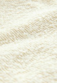 Texturiertes cremefarbenes Material mit einer weichen, plüschigen Oberfläche. Enthält eine Mischung aus erhöhten und flachen Bereichen, die ein einladendes haptisches Erlebnis schaffen.