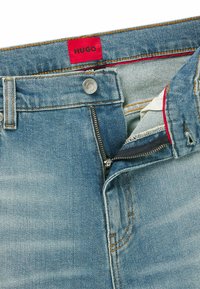 Jeans in denim azzurro chiaro con zip e bottone, etichetta rossa "HUGO" e cuciture a contrasto. Texture liscia con design classico delle tasche.
