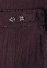Tissu violet foncé avec deux boutons gris à quatre trous sur un rabat de ceinture réglable cousu dans le vêtement.