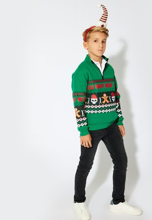 Polaire verde con un cuello con cremallera que presenta patrones festivos y texto. Combinado con jeans negros y zapatillas blancas. Diadema a rayas rojas y blancas.