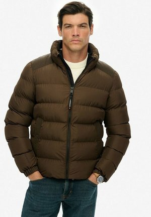 Hombre con chaqueta acolchada marrón y capucha, suéter blanco, jeans azules y reloj, de pie con las manos en los bolsillos contra un fondo blanco.