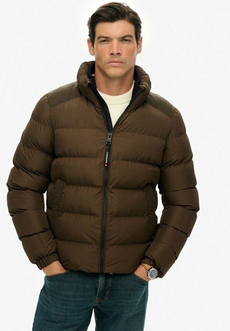 Mand iført brun polstret jakke med hætte, hvid sweater, blå jeans og armbåndsur, stående med hænderne i lommerne mod en hvid baggrund.