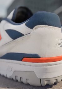 Sneaker bianca con dettagli blu navy e una striscia arancione. Materiali in suede testurizzato e mesh sulla parte superiore. Suola in gomma resistente con scanalature.