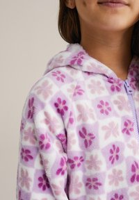 Polár kapucnis dzseki lila és rózsaszín árnyalataiban virágmintával, elöl cipzárral és puha, texturált anyagból.