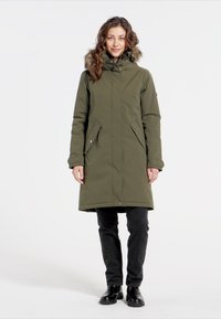 Olivengrøn isoleret parka med en pelsforet hætte, frontlommer, lange ærmer og en mellemlang længde, båret over sorte bukser og støvler.