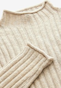 Beige gestrickter Pullover mit hohem Kragen, der über vertikale Rippen und eine weiche Textur verfügt. Die Ärmelbündchen haben ein umgeschlagenes Kanten-Detail.