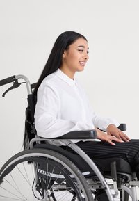 Rollstuhl mit einem silbernen Rahmen und schwarzem Sitz, besetzt von einer Person, die ein weißes Hemd mit Knöpfen und schwarze gestreifte Hosen trägt, mit herabfallendem Haar.