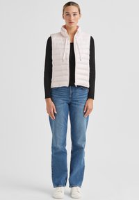 Hellrosa gesteppte Weste mit hohem Kragen, Reißverschluss und schwarzem, geripptem Langarmshirt, kombiniert mit blauen Jeans und weißen Turnschuhen.