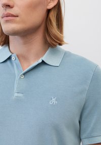 Himmelblaues Polohemd aus strukturiertem Stoff, mit Kragen, zwei Knöpfen und einem kleinen weißen gestickten Logo auf der Brust.