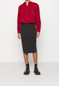 Red long-sleeve button-up shirt with white logo pattern, paired with a black knee-length skirt featuring side stripes, worn with black ankle boots.