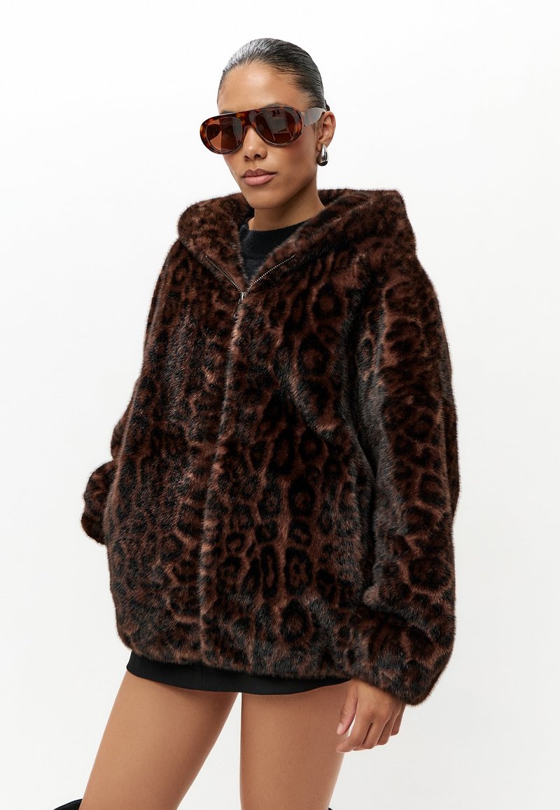 Frau trägt große Sonnenbrille mit Schildpattfassung und eine dunkelbraune Leopardenmuster Kunstpelzjacke mit Kapuze.