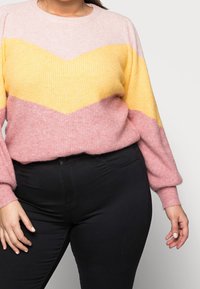 Kvinna som bär en pastellfärgad tröja i rosa, gul och ljusrosa färgblock tillsammans med svarta byxor, stående mot en enfärgad bakgrund.